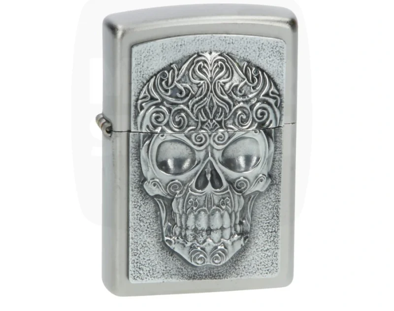 Briquet Zippo argenté brossé avec plaque en relief d'un crâne richement décoré de motifs ornementaux.